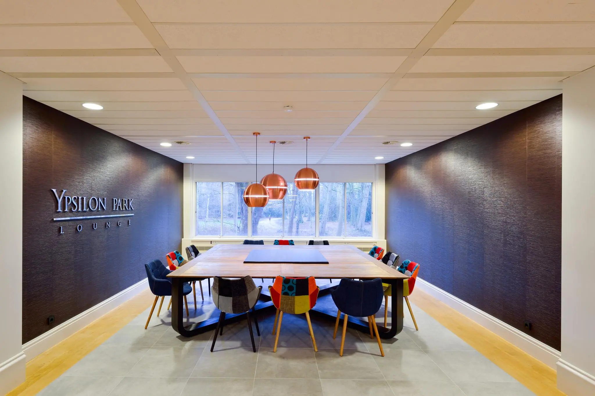 Vergaderruimte in Ypsilon Park Lounge met een grote houten tafel, kleurrijke stoelen en drie koperen hanglampen.